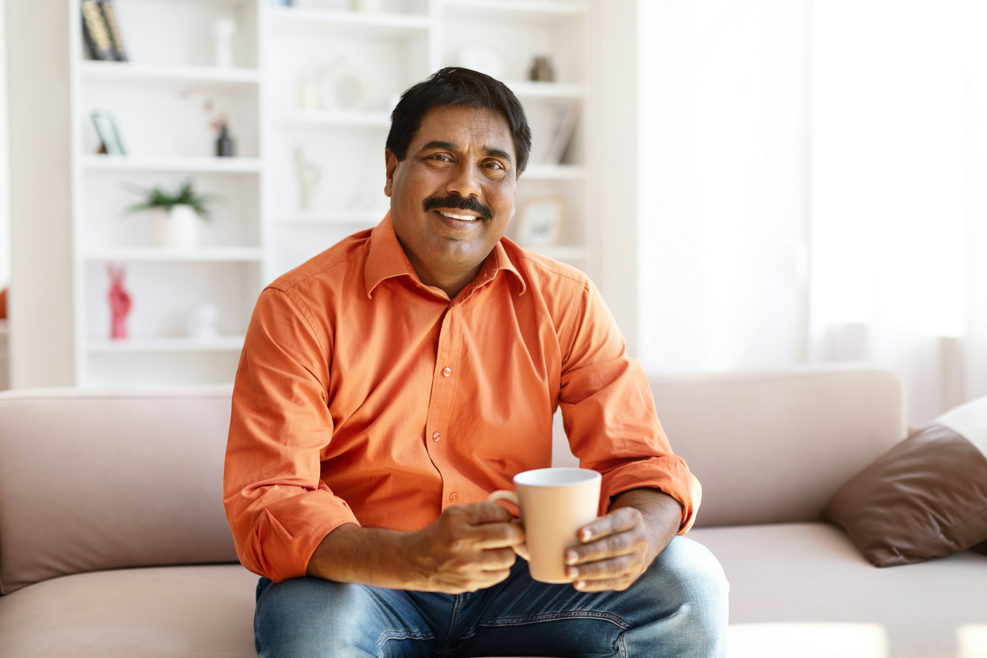 Happy mature indian man sitting on couch, drinking coffee