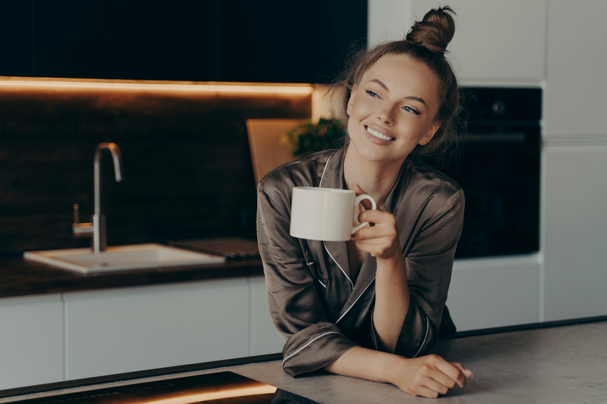 Happy smiling young woman in brown satin pajama enjoying aromatic morning coffee in kitchen